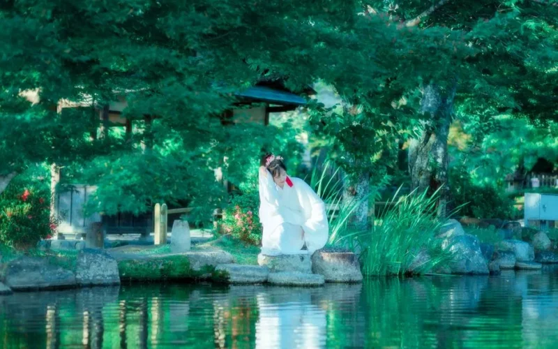 Woman in traditional kimono sitting by a peaceful lake in a lush Japanese garden.