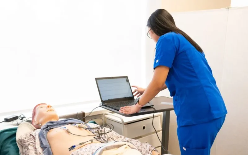 Nurse in training using a laptop with a medical dummy in a clinical simulation lab.