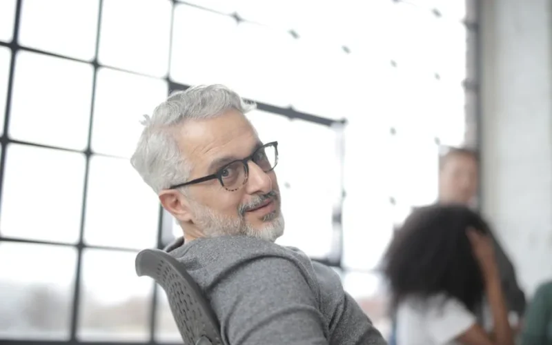 Positive gray haired bearded male manager wearing eyeglasses sitting in computer chair while working in office against big window near group of diverse employees and looking over shoulder