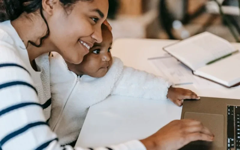 Side view of crop cheerful young black mother with curious infant in warm casual clothes using laptop sitting at table at home