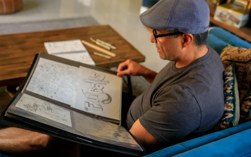 A man sits on a sofa reviewing his art portfolio filled with various illustrations.