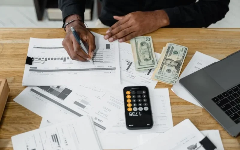 Man working on financial reports with calculator, money, and laptop on a desk.