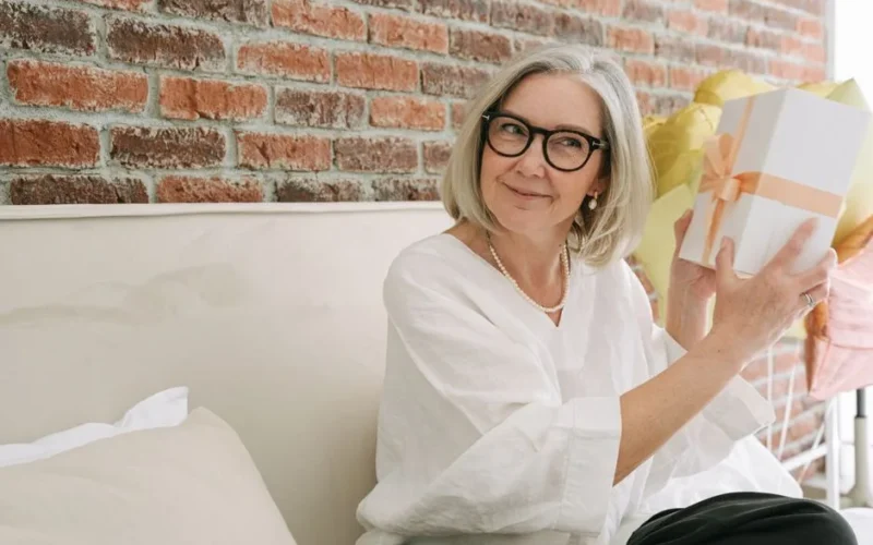Senior woman in white blouse happily holding a gift box indoors in front of a brick wall.