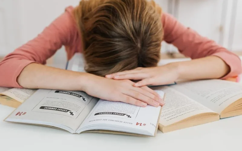 A student overwhelmed by studying falls asleep on open books at a desk, representing academic fatigue.