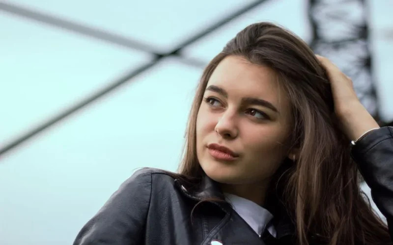 A young woman with long hair in a leather jacket looking over her shoulder outdoors.