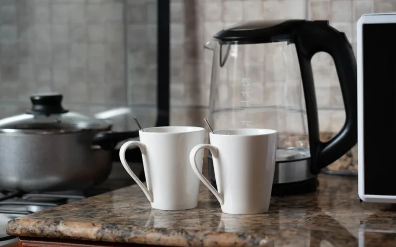 A warm indoor shot of two coffee mugs next to a kettle on a stylish kitchen counter.