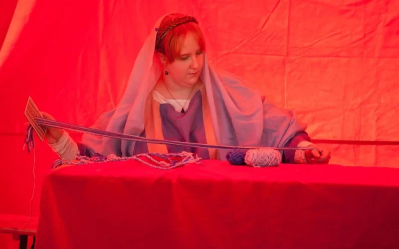 Woman in historical attire weaving with loom, red tent background, reenactment event.