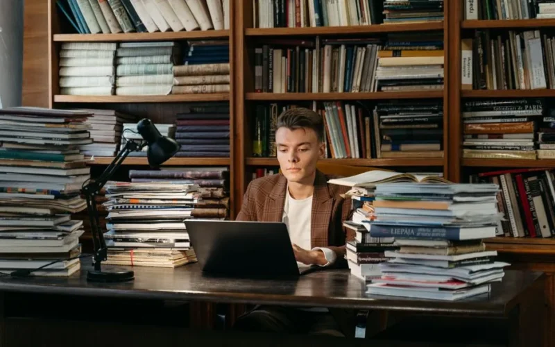 A young man intensively studying at a library surrounded by books, using a laptop.