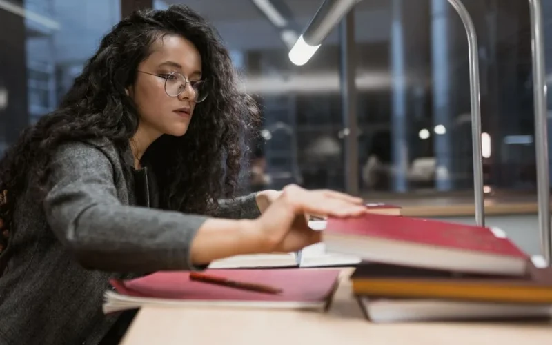 A woman with glasses studying at a desk under a lamp in a library at night.