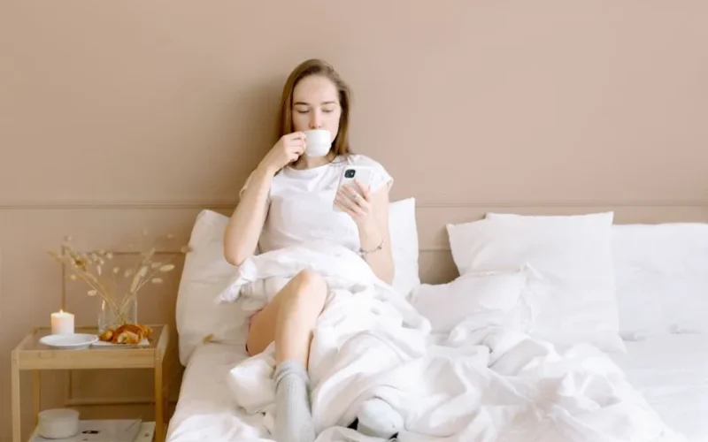 Relaxed woman in bed drinking coffee and using smartphone in a cozy setting.