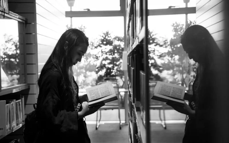 A woman engrossed in a book beside a reflective window in a library.