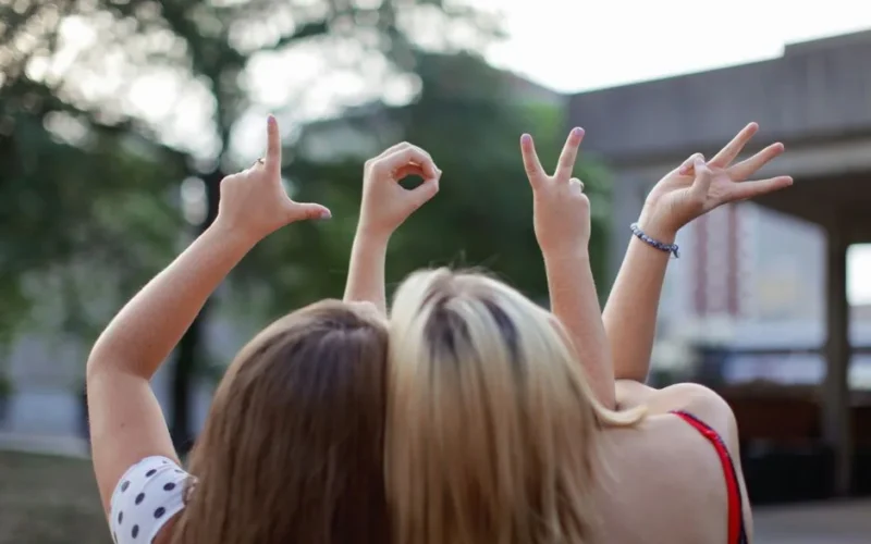 Two young women forming the word 'LOVE' with hand signs, symbolizing friendship in a summer setting.