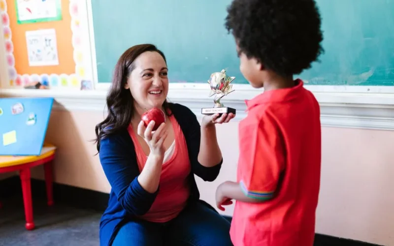 A teacher holding an apple and a student holding a trophy in a classroom setting.