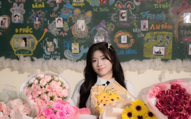 A young woman poses with vibrant bouquets of flowers against a colorful classroom blackboard.