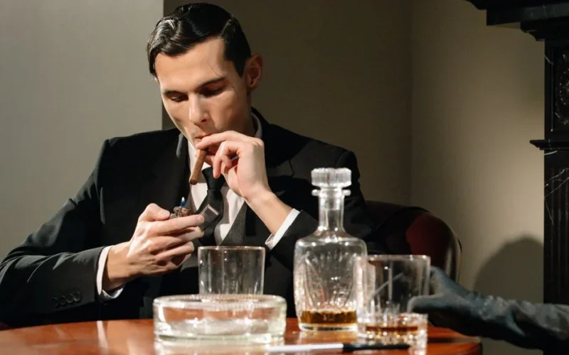 A sophisticated man in a suit smokes a cigar while enjoying a drink, exuding luxury and vintage style.