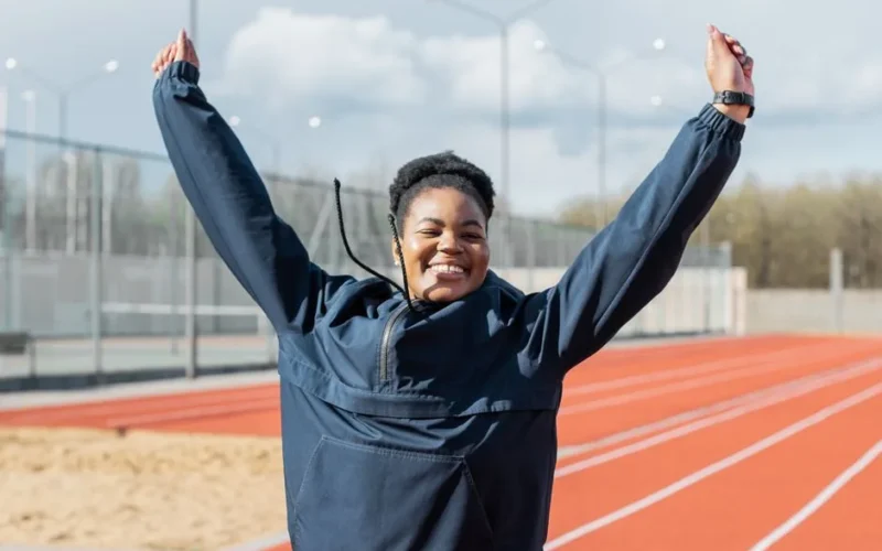 Smiling athlete in activewear celebrating victory with raised arms on outdoor track.