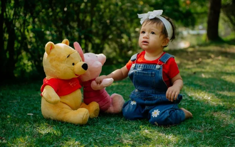 Adorable child in denim and red plays with Pooh Bear plush toys on grass.