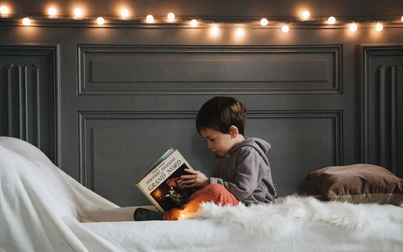 A young boy reads a book in a cozy bedroom setting with warm string lights and soft textures.