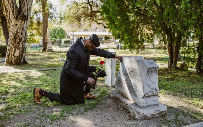 A man kneels at a gravestone, holding a red rose, mourning in a peaceful cemetery.