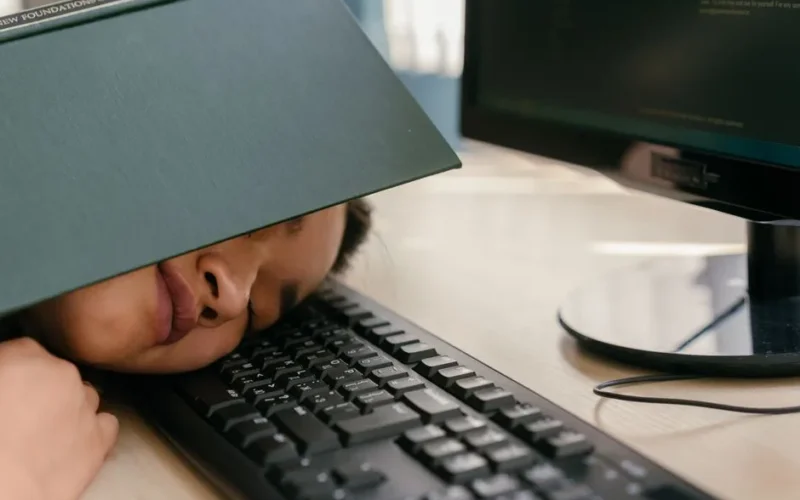 A young woman falls asleep on a keyboard with a textbook on her face, depicting study fatigue.