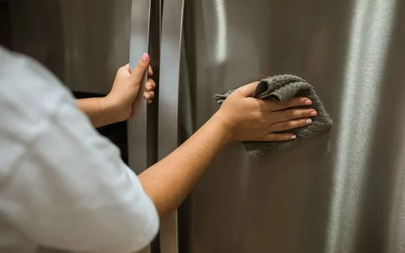 Close-up view of hands cleaning a stainless steel refrigerator with a cloth indoors.