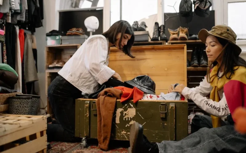 Two women sorting through vintage clothing in a thrift shop, surrounded by shoes and hats.