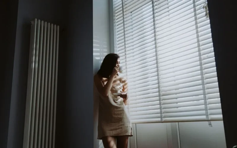 A woman in soft morning light stands by the window, holding a cup and saucer, savoring a peaceful moment.