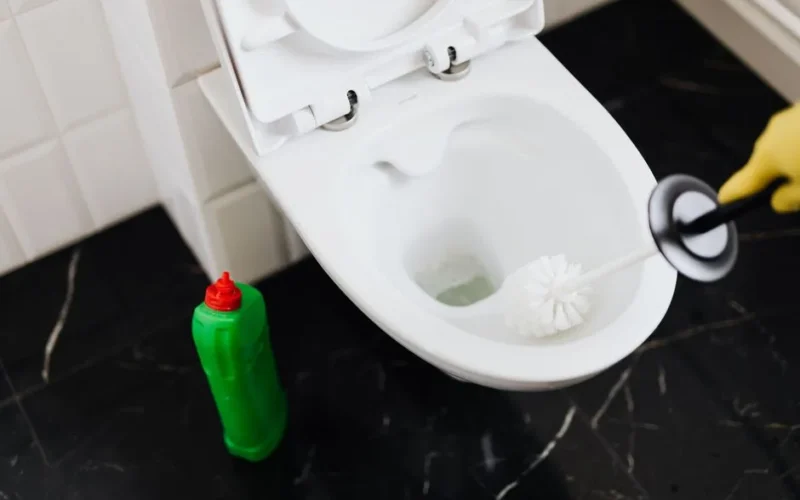From above of crop anonymous person in rubber gloves using liquid toilet sanitizer and brush to clean toilet bowl in bathroom