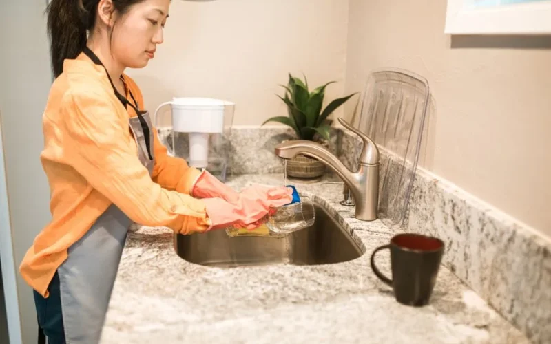 A woman wearing gloves washes a drinking glass at a kitchen sink, focusing on hygiene and cleanliness.