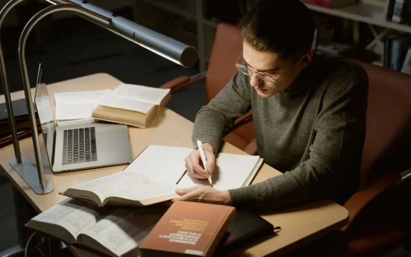 A young adult studying with books and a laptop, focused on taking notes at a desk under a lamp.