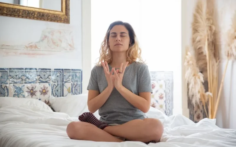 A woman meditating in a peaceful bedroom setting with soft natural light.
