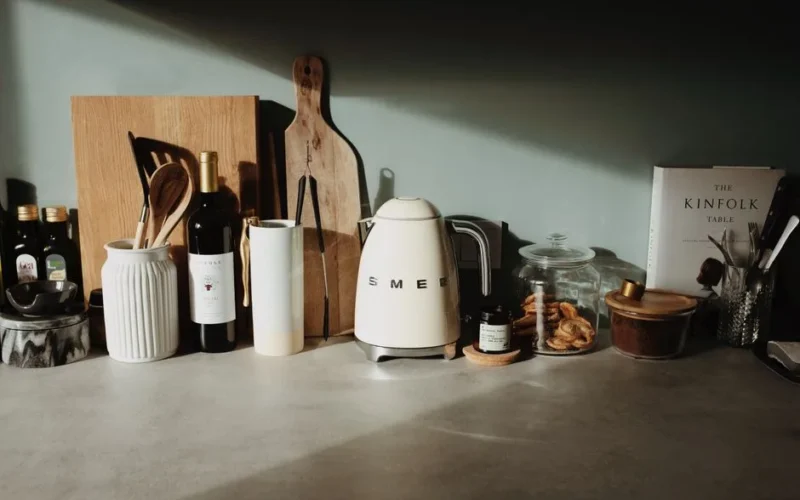 Various glass and ceramic bowls stylish electric kettle cutting boards and kitchen utensils locating on stone counter against gray wall in cozy kitchen