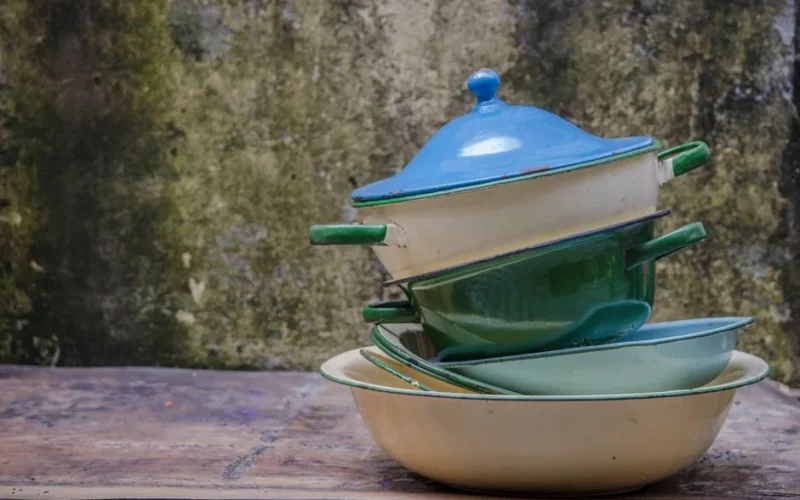 A rustic stack of colorful vintage enamel pots and pans outdoors against a textured wall.