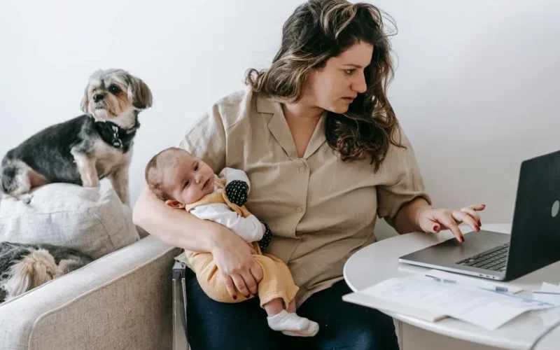 Mother balancing work from home with parenting, holding baby and typing on laptop.