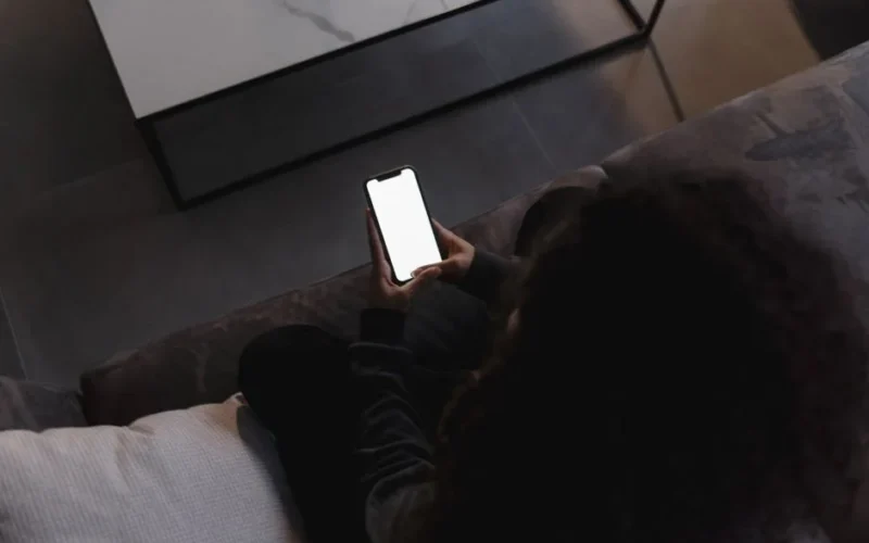 A person holding a smartphone with a blank screen, seated indoors on a couch.