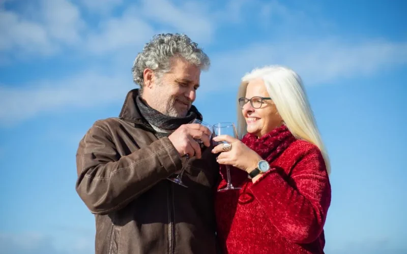 Elderly couple sharing glasses of wine, embracing love and happiness under a clear blue sky.