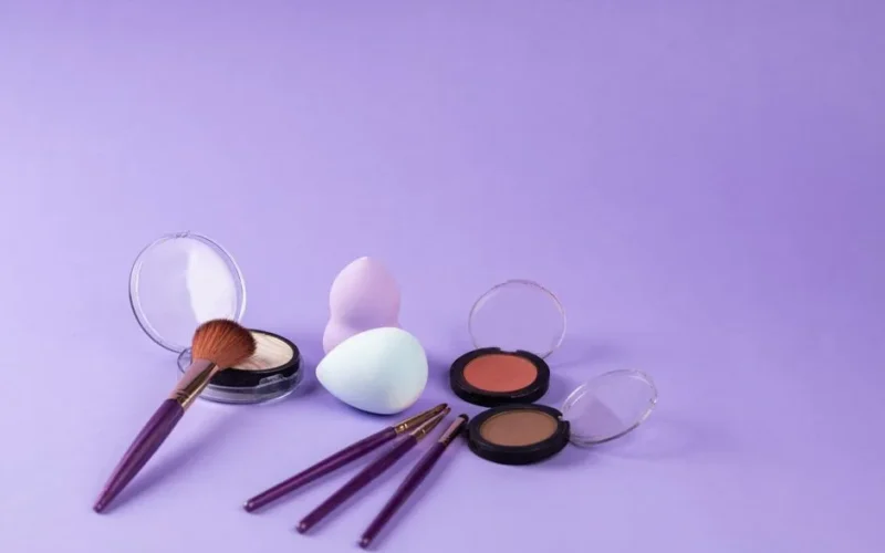 A colorful arrangement of makeup products including brushes and beauty sponges on a purple background.