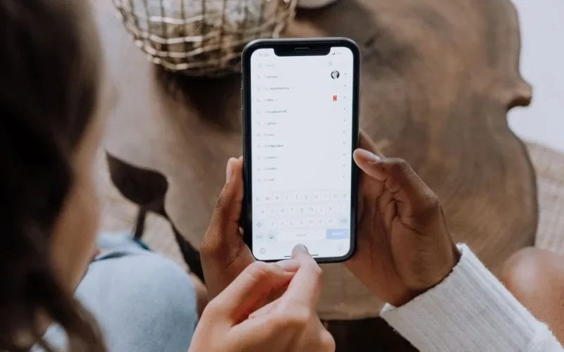 Close-up of hands holding a smartphone, searching for apps like TikTok on a wooden table.