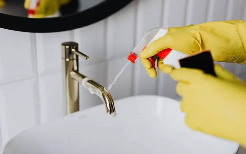 From above anonymous housekeeper in yellow rubber gloves spraying cleaner to chrome water faucet in bathroom