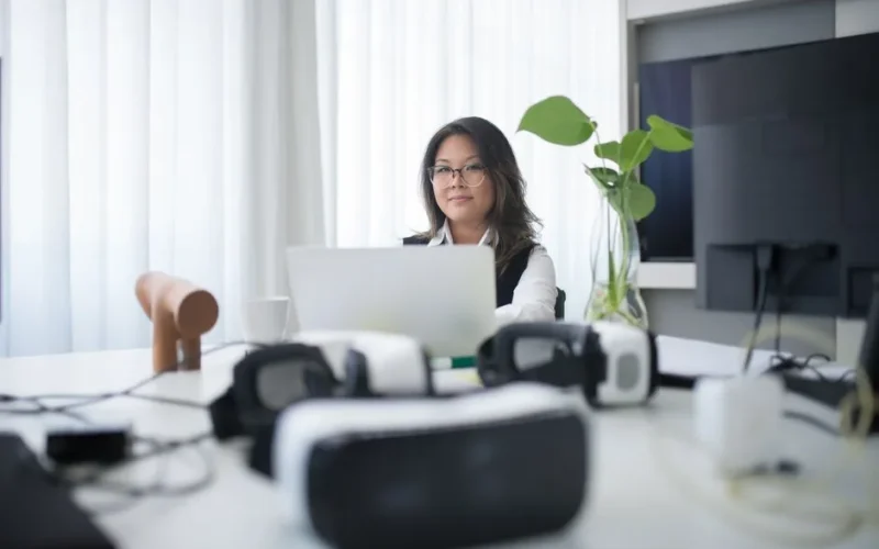 Asian woman in office using laptop with VR headsets on table, symbolizing modern workspace.