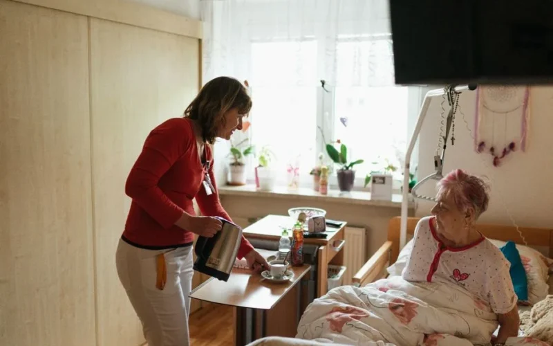 A caregiver serves tea to a senior woman in a cozy bedroom setting, fostering comfort and care.