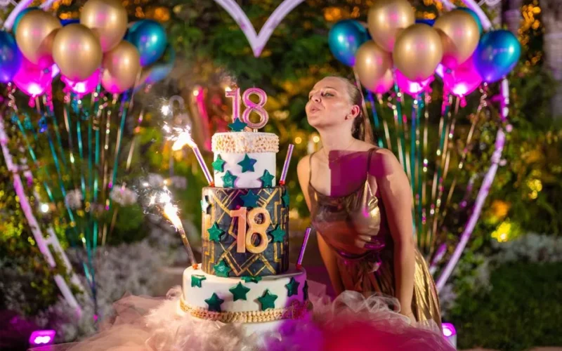 Content young female in elegant dress blowing shiny candles on birthday cake while standing with pouting lips near decorative heart and colorful balloons during party
