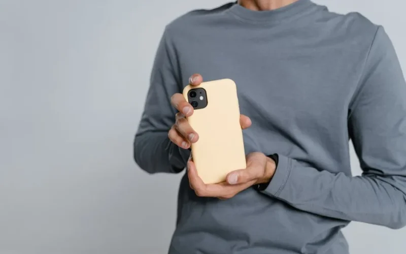 Close-up of a man holding a smartphone with a yellow case, wearing casual attire, shot in a studio.